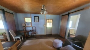 A compact living room with a wooden ceiling, a ceiling fan, a dining table with chairs, a small ottoman, and a sofa.