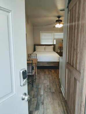 A bedroom with a wooden floor, a bed with white bedding, a ceiling fan, and a window with blinds.