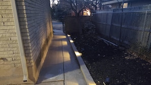 Een smal betonnen pad dat leidt naar een gebouw van baksteen, met een metalen hek aan één kant.