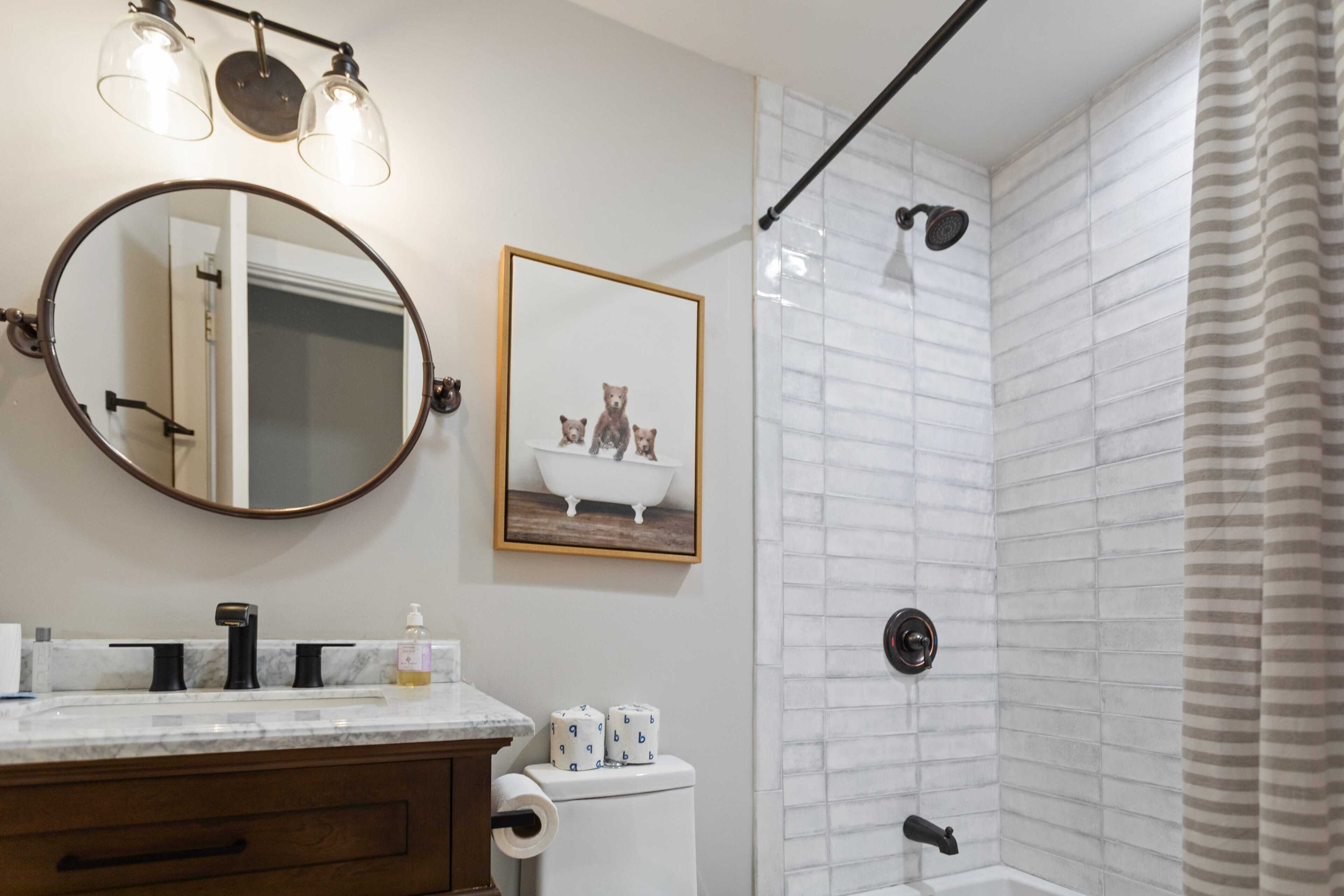 A bathroom with a round mirror, a framed picture of bears, a toilet paper holder, and a shower with a towel rack.