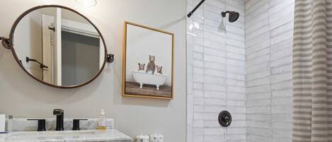 A bathroom with a round mirror, a framed picture of bears, a toilet paper holder, and a shower with a towel rack.