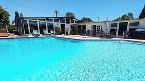 Una piscina amb gandules i ombrelles, envoltada d'un edifici modern amb grans finestres i una zona de terrassa coberta.