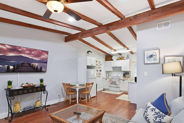 A spacious living room with a wooden ceiling, a dining area with chairs and a table, a kitchen with white cabinets, and a large painting on the wall.