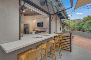 An outdoor dining area with a long table and multiple chairs, a kitchen in the background, and a view of the garden.