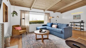 Ein Wohnzimmer mit einem blauen Sofa, einem Holztisch, einem roten Sessel und einem großen Fenster mit Vorhängen.