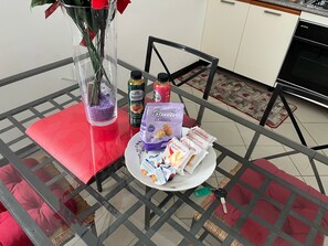 A glass table with a red cushion, a vase with a red flower, and various food items including a plate of snacks and two bottles. - Nice Apartment With sea View in Genoa (Genoa)