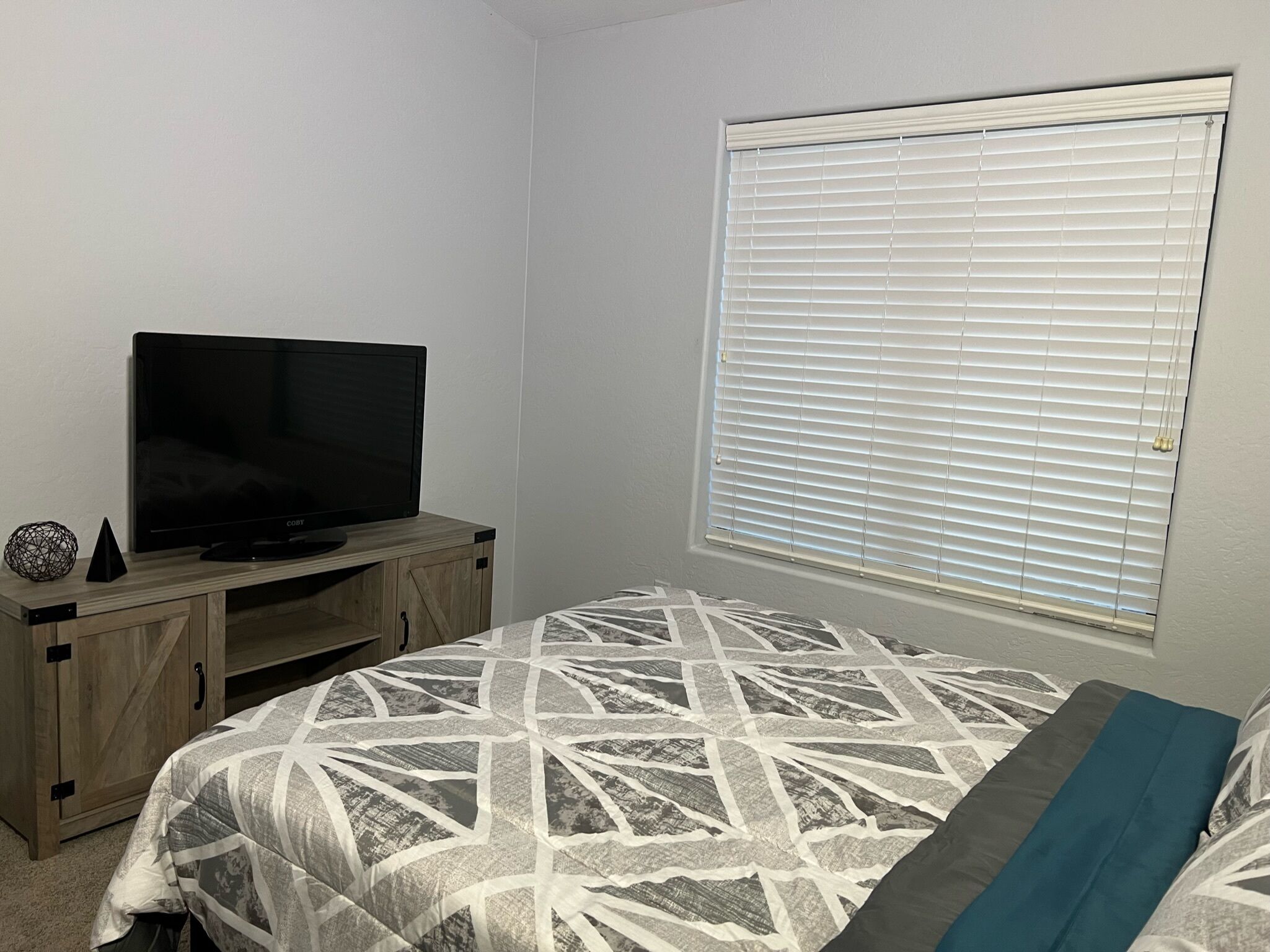 A bedroom with a bed, a TV on a wooden stand, and a window with blinds.