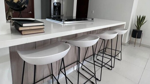 Una cocina moderna con encimera blanca, campana extractora de acero inoxidable y tres taburetes con estructura de metal negro.