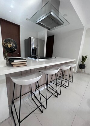 Une cuisine moderne dotée d’un plan de travail blanc, d’une hotte en acier inoxydable et de trois tabourets de bar à structure en métal noir.