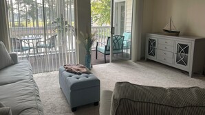 Sala de estar com sofá, puf e aparador com gavetas. Há uma porta de correr que dá para uma área externa com mesa e cadeiras.