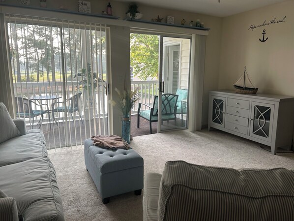 A living room with a sofa, ottoman, and a cabinet with drawers. There is a sliding glass door leading to an outdoor area with a table and chairs.
