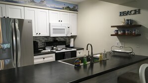 Cozinha com armários brancos, geladeira de aço inoxidável, micro-ondas e fogão. Há uma pia com torneira, um banquinho de bar e uma prateleira com objetos decorativos.