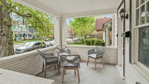 A covered porch with wicker chairs and a concrete table, overlooking a street with parked cars and houses. - ⭐Modern 2 Bedroom Unit ⭐ Convenient ⭐ Near Downtown, Convention & Hospitals ⭐ (Indianapolis)