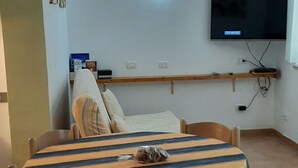 Un comedor con mantel a rayas, sillas de madera, un televisor montado en la pared y una estantería con objetos.