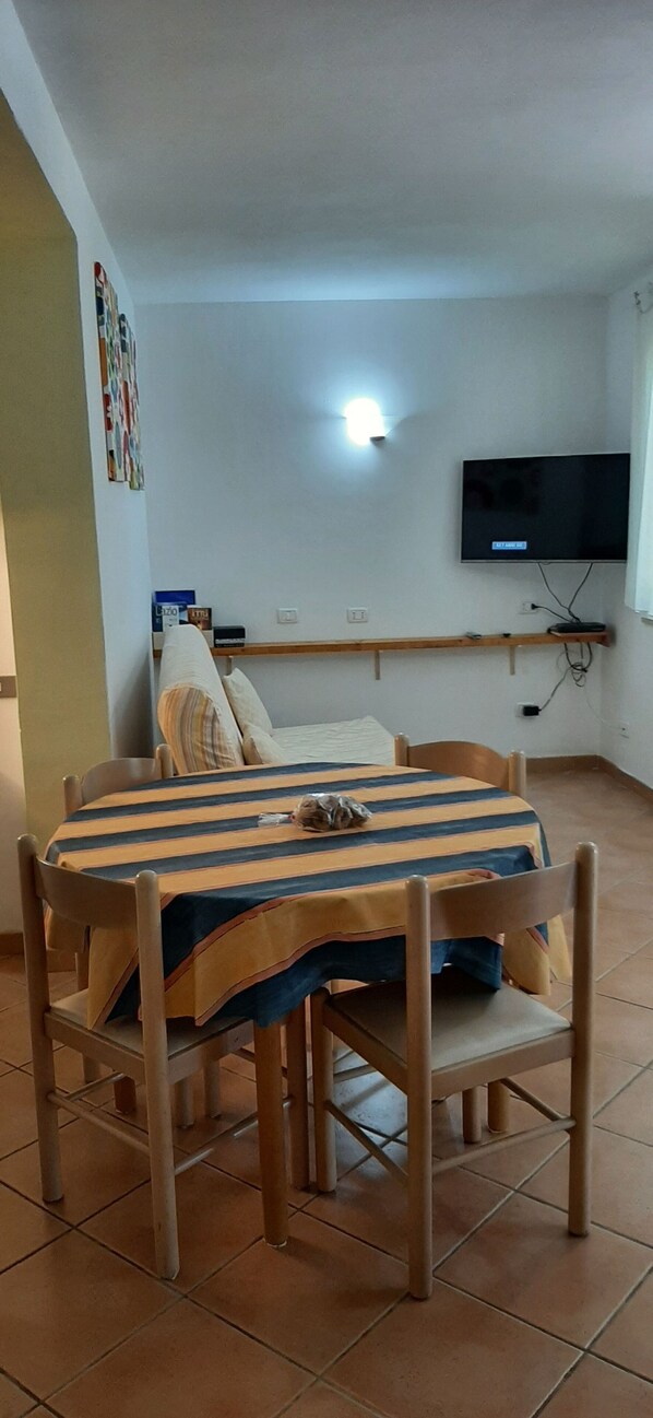 A dining area with a striped tablecloth, wooden chairs, a television mounted on the wall, and a shelf with items.