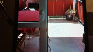 Ein Zimmer mit einem roten Bett, einem Sessel und einem kleinen Tisch. Es gibt einen Balkon mit roter Brüstung und einem Blumentopf.