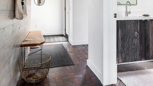Un couloir doté d’un plancher en bois, d’un banc, d’un panier, d’un miroir et d’une porte donnant sur une salle de bain équipée d’un lavabo et d’un miroir.