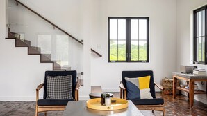 Un salon moderne avec un escalier, deux fauteuils, une table basse et une grande fenêtre donnant sur un paysage verdoyant.