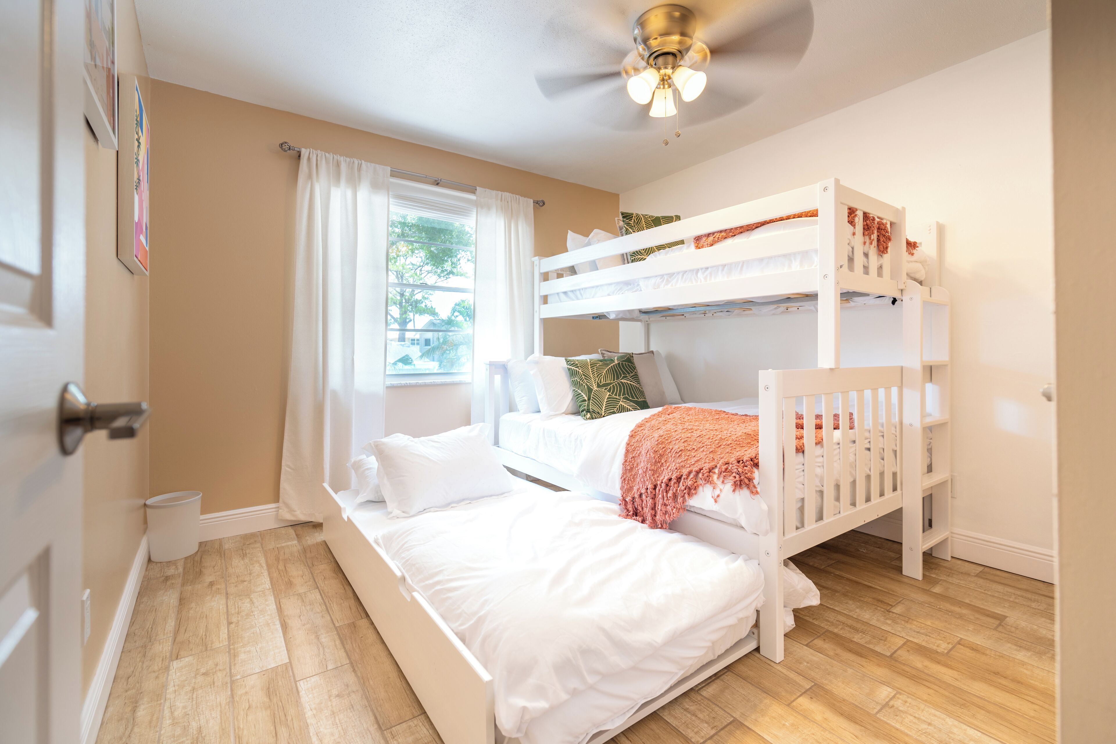 Un dormitorio con una cama litera, una cama individual, un ventilador de techo, una ventana con cortinas y vistas al exterior.