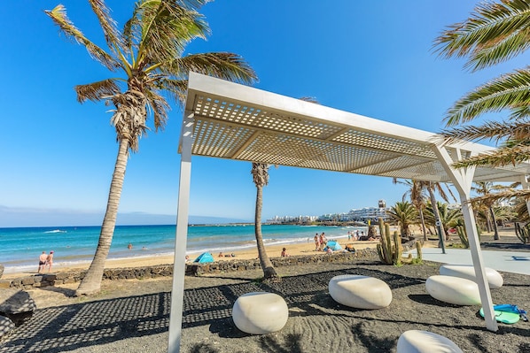 A beach with a white pergola, palm trees, and a clear blue sky. - Lanzahost "Casa Juan" - Playa Bastian (Costa Teguise)