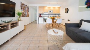 Una sala moderna con un sofá, una mesa de centro y un televisor. Se observa una cocina al fondo con armarios de madera y una zona de comedor con sillas.