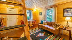 Ein gemütliches Blockhaus-Interieur mit Holzbalkendecke, einer Couch mit Kissen, einem kleinen Tisch mit Lampe und einem Fenster mit Vorhängen.