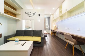 Un salon moderne avec un canapé, une table basse et un espace bureau. Un mur porte un message positif et une fenêtre est agrémentée de rideaux.
