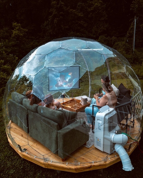 A transparent dome structure with a wooden deck, seating, and a television.