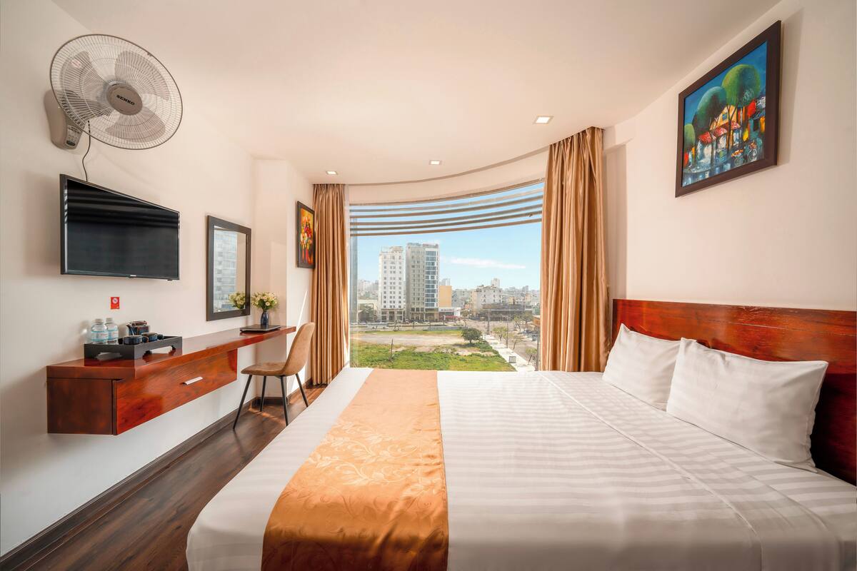 Une chambre d’hôtel avec un lit, un bureau et une chaise, une télévision, un ventilateur et un tableau.