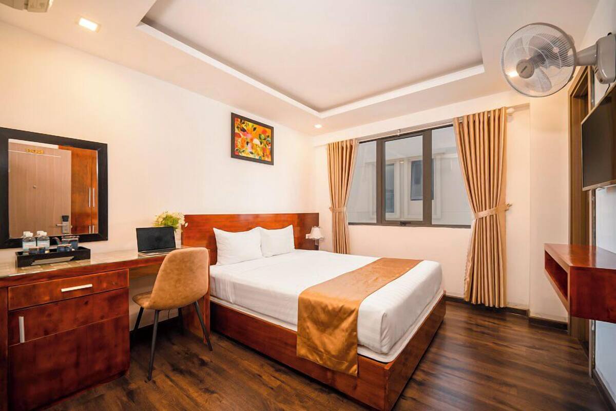 Une chambre d’hôtel avec un lit en bois, un bureau avec une chaise, une grande fenêtre avec des rideaux, un ventilateur de plafond et un tableau accroché au mur.