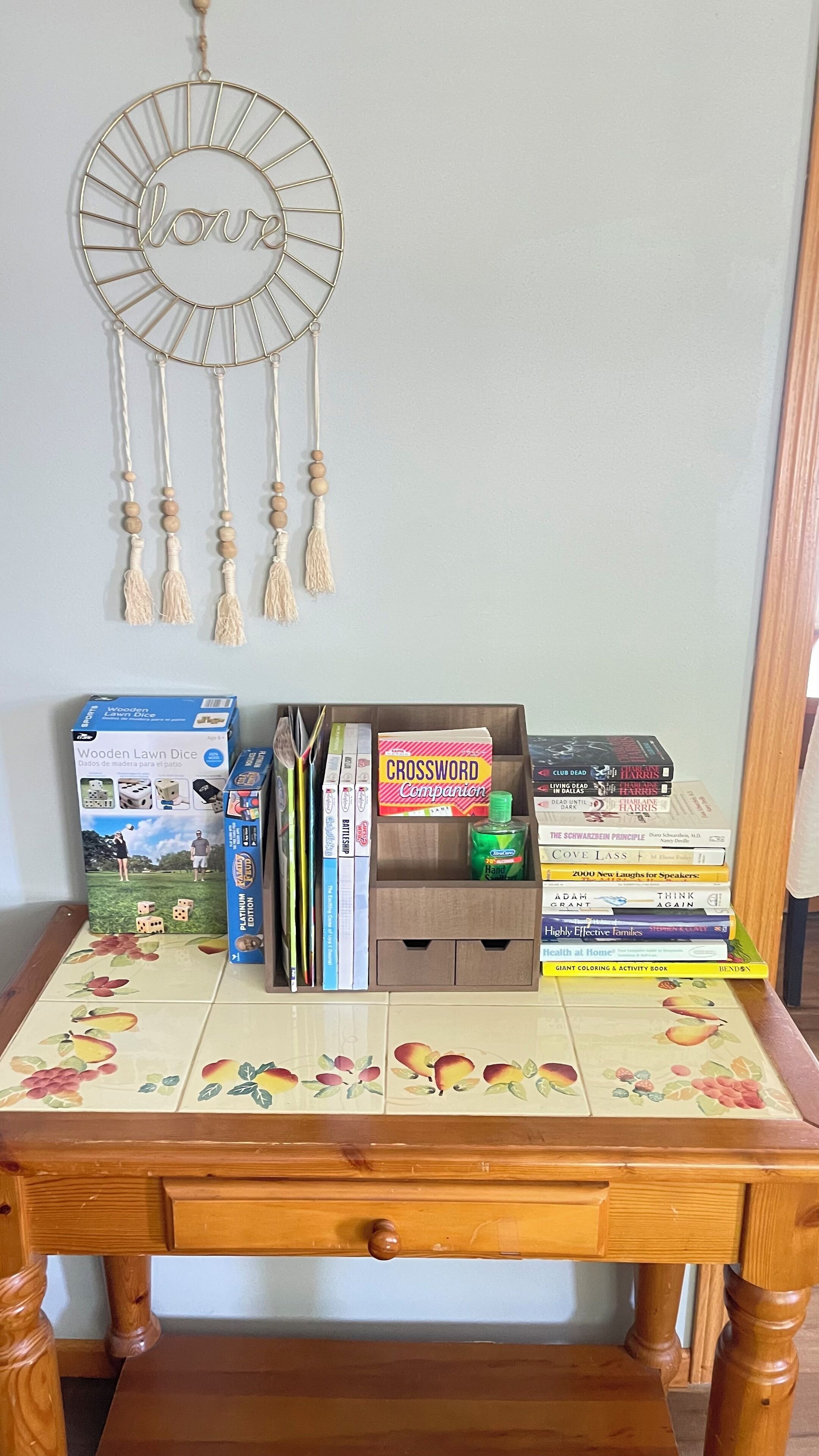A wooden table with a floral tile design, a dreamcatcher, a wooden lawn dice, a crossword companion box, and a stack of books.