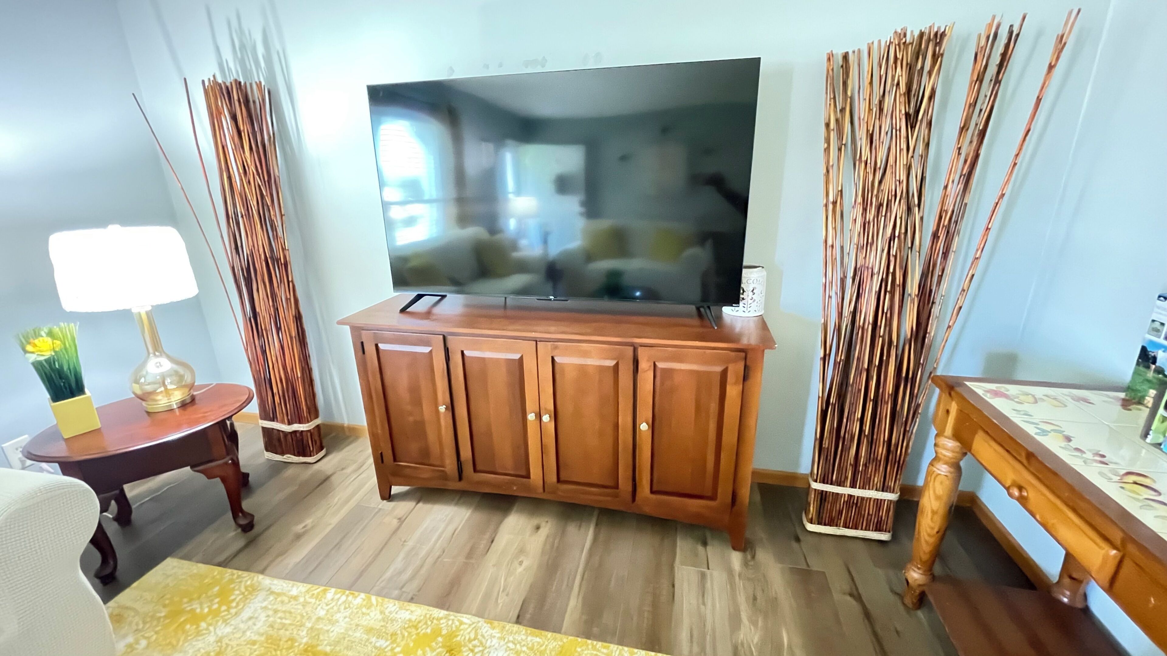 Una sala de estar con un aparador de madera, un televisor de pantalla plana, una mesita con una lámpara y estructuras decorativas que parecen cañas.