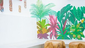 A room with a white wall, a colorful tropical plant mural, and two wooden stools.
