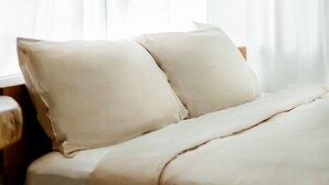 A bed with white bedding and pillows in a room with a wooden headboard and a window with sheer white curtains.