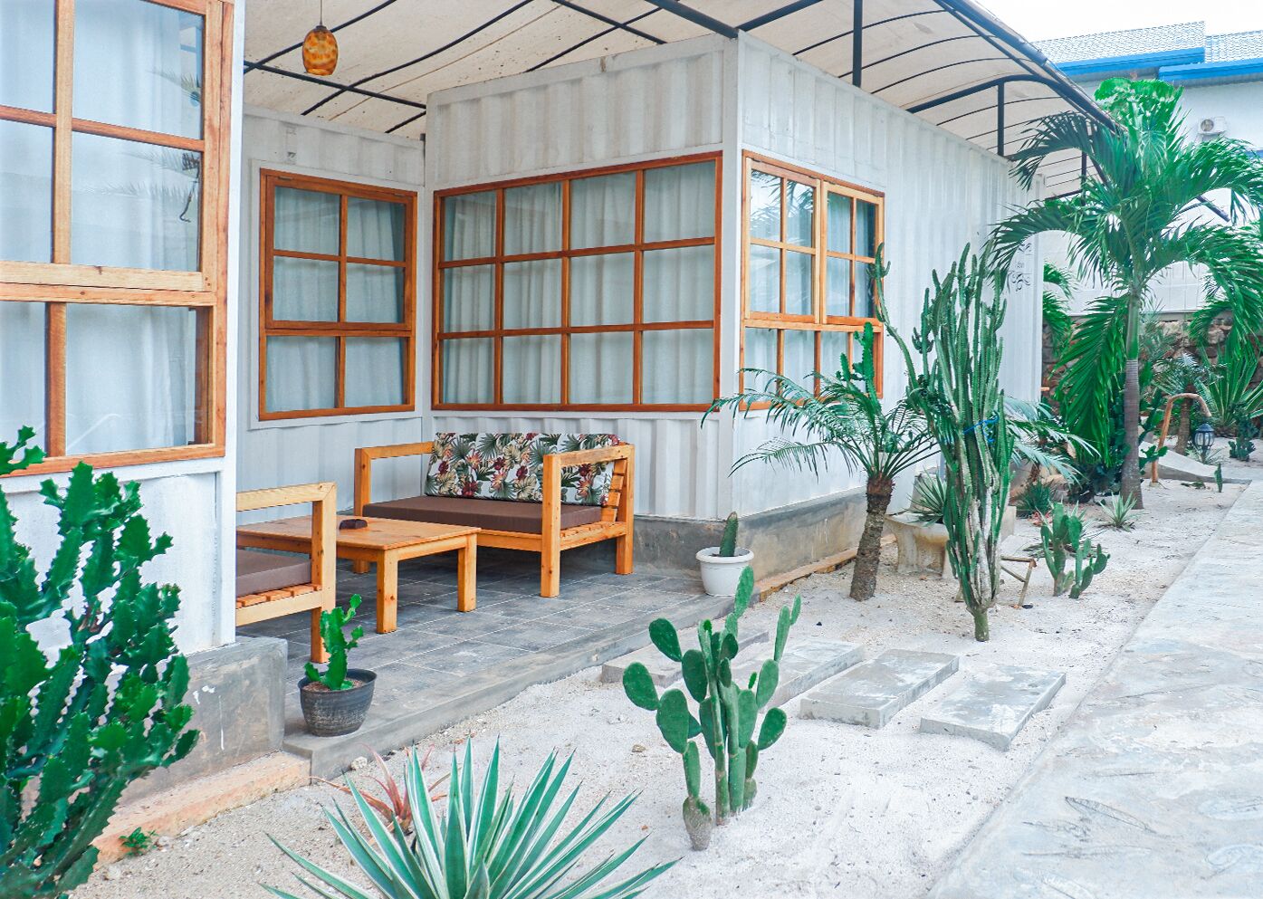 A covered outdoor seating area with a wooden bench and table, surrounded by potted cacti and other plants.