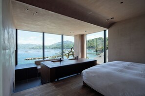A modern bedroom with a large bed, a wooden desk with a sink, and a view of a lake and mountains.