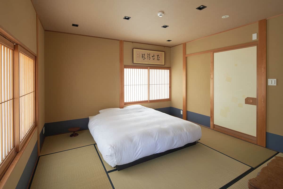 Ett traditionellt japanskt rum med tatamimattor på golvet, en säng, en skjutdörr och ett fönster med vertikala persienner.