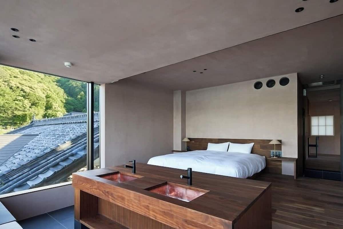 Ett modernt hotellrum med ett stort fönster, en träinredd sminkbord med handfat, en säng med vita sängkläder och ett träfärgat sänggavel.