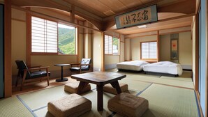 Una habitación tradicional japonesa con suelo de tatami, una mesa baja, dos futones de tatami para sentarse, una cama y vistas a la montaña a través de amplias ventanas.