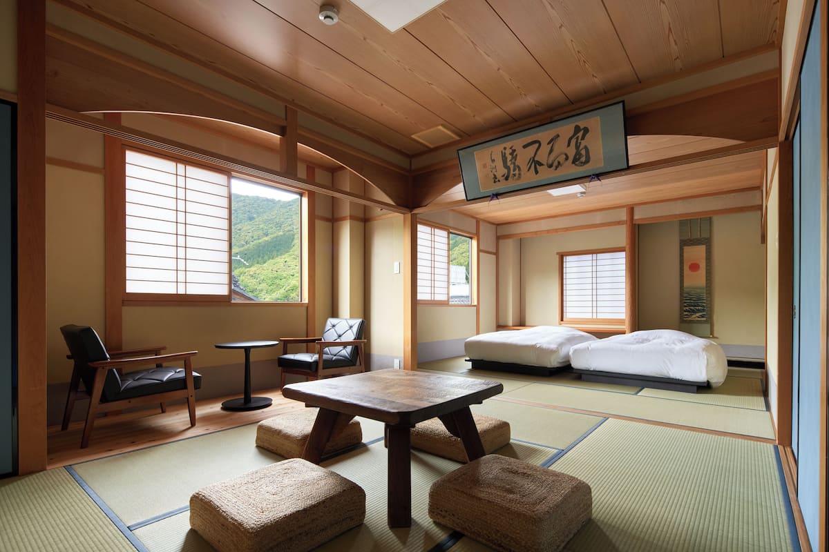 Ett traditionellt japanskt rum med tatamimattor på golvet, ett lågt bord, två tatamimattor som används som sittplatser, en säng och en utsikt över ett berg genom stora fönster.