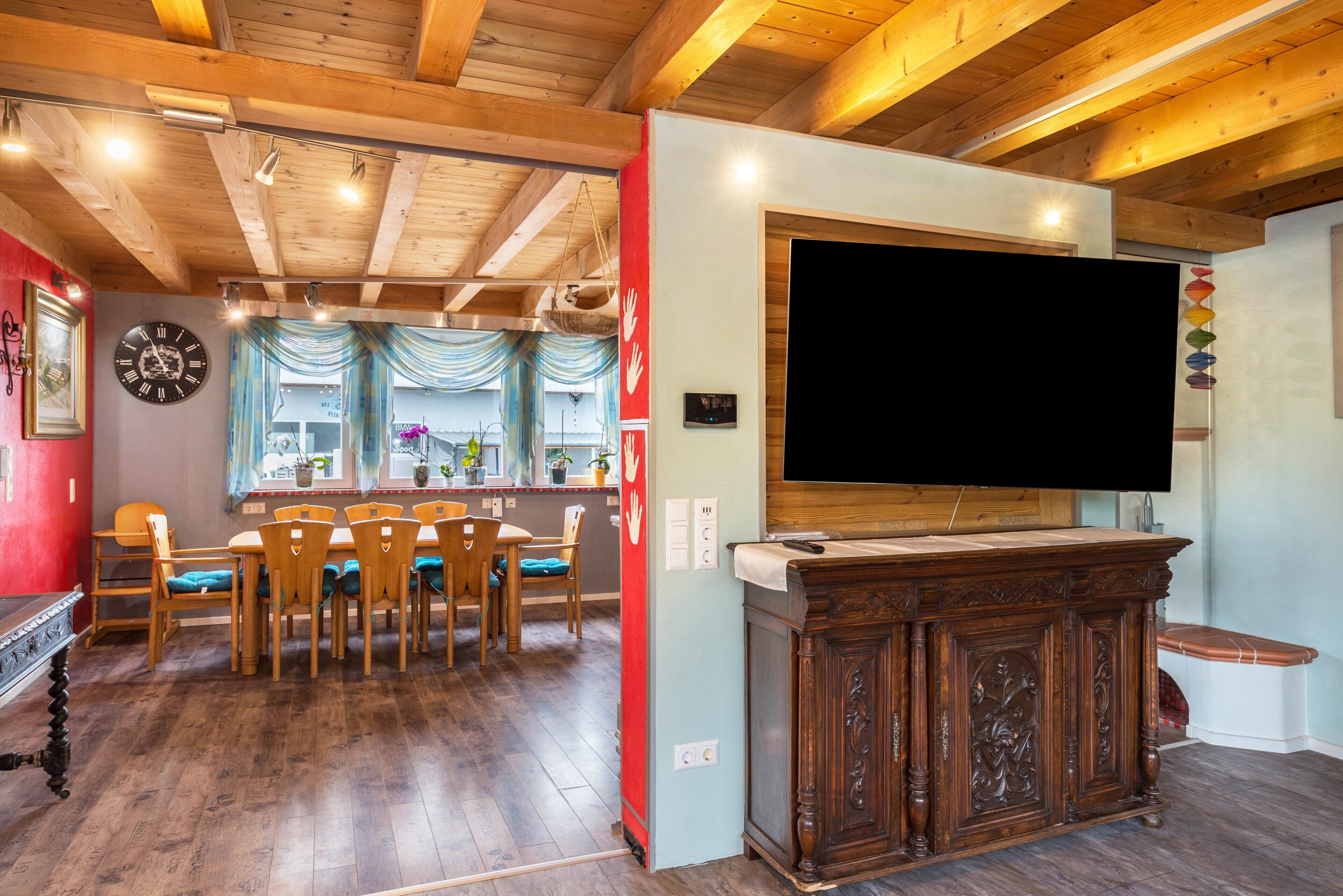 Zona pranzo con sedie di legno e un tavolo, una TV su un mobile in legno e una sveglia appesa al muro.