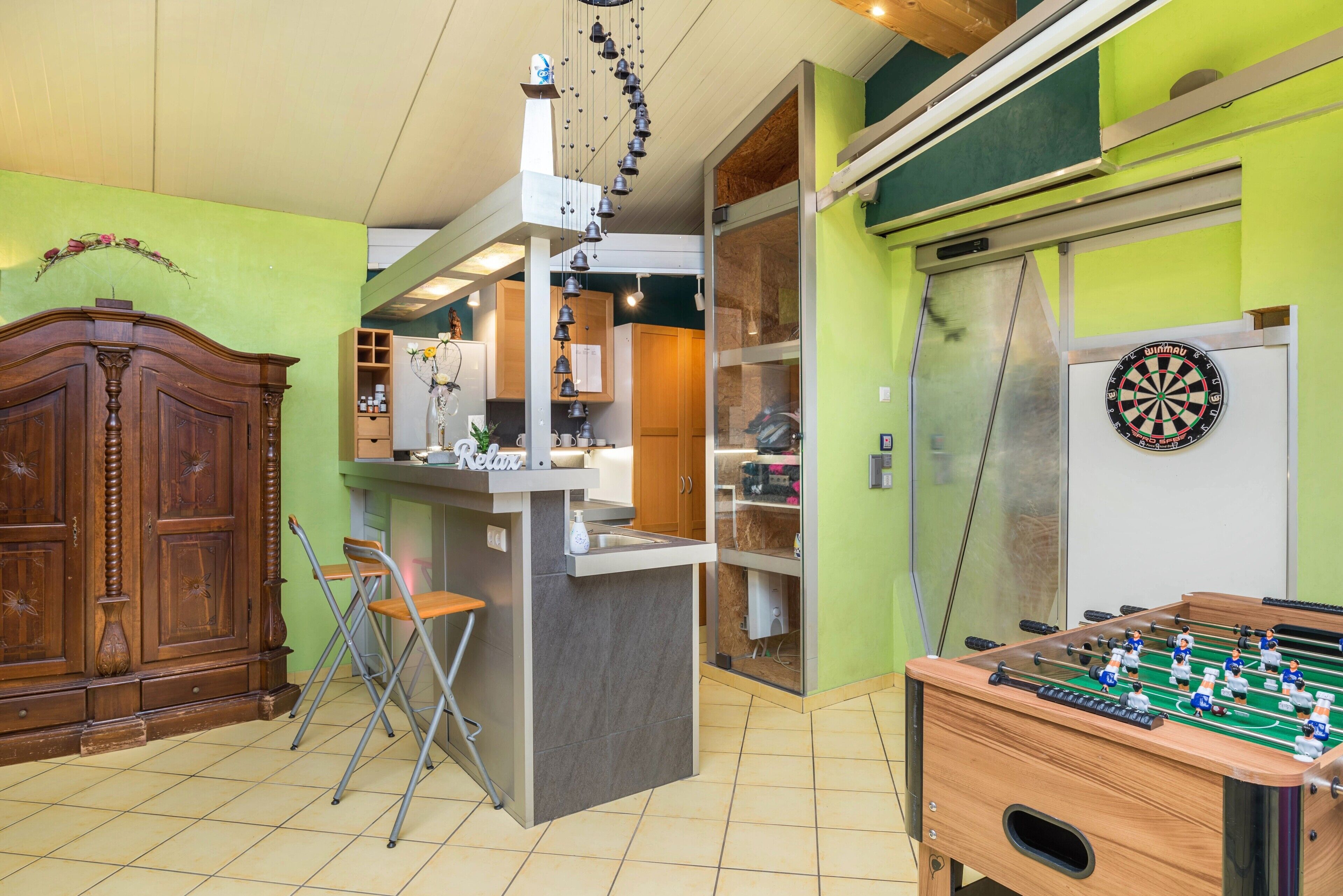 Ein Zimmer mit Holzschrank, Kicker, Dartscheibe und Küchenzeile mit Regalen und Kühlschrank.