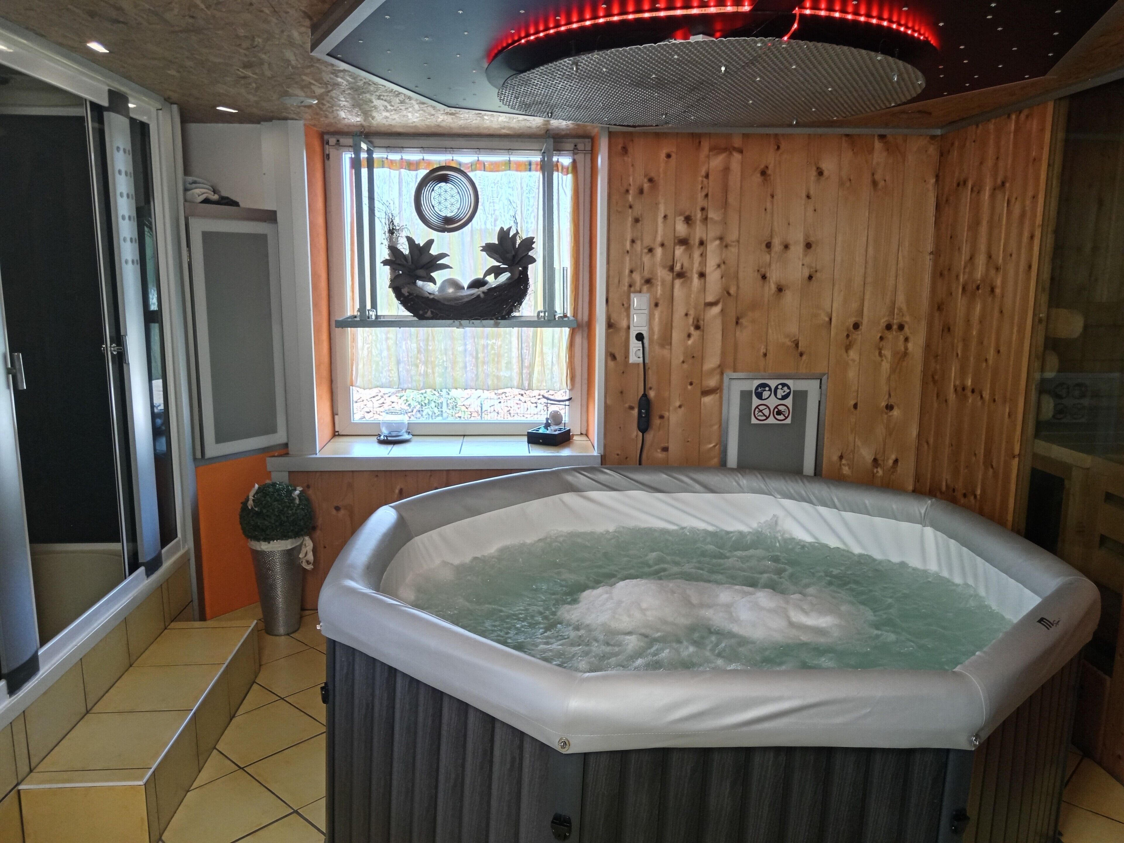 A hot tub in a room with wooden walls, a window with a decorative item, and a ceiling light fixture.
