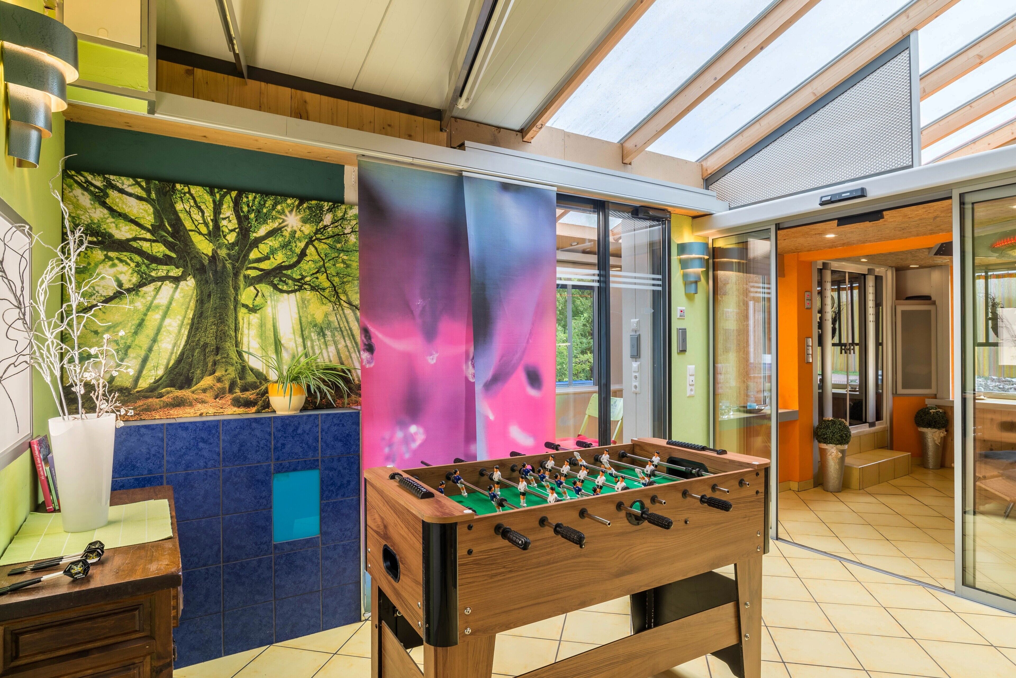 Una habitación con mesa de fútbol, un ventanal grande y un mural de un árbol.
