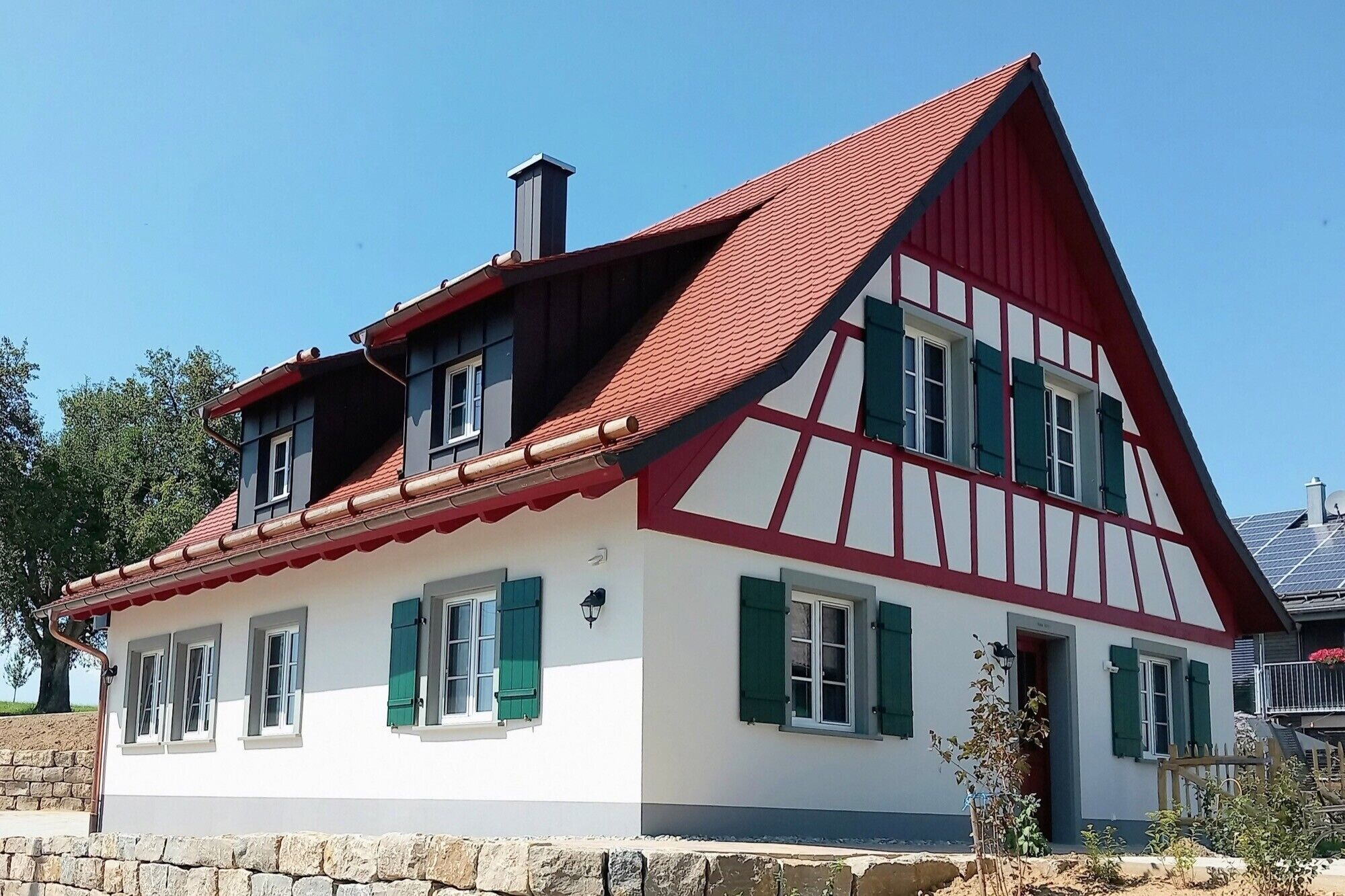 Une maison à deux étages, avec un toit rouge à forte pente, des murs blancs et des volets verts.