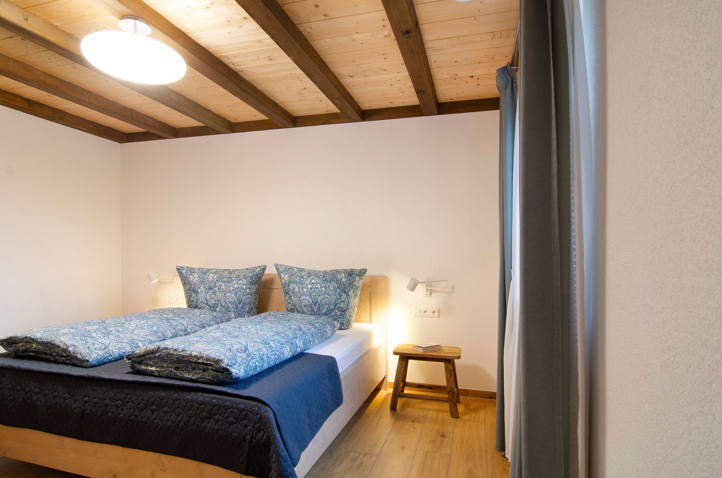 Een slaapkamer met een bed, nachtlampen, een klein houten krukje en een plafondlamp.