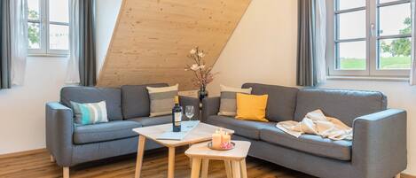 Ein modernes Wohnzimmer mit schrägverdeckter Decke, einem grauen Sofa, einem kleinen Holztisch und einem Fenster mit Vorhängen.
