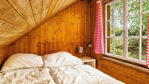 Una habitación de cabaña de madera con una cama, una mesita de noche, una lámpara y una ventana con cortina de cuadros rojos y blancos.
