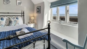 A bedroom with a bed, a nightstand, a lamp, a window with a view of the sea, and a chair. - Captain's Cottage, Minehead (Minehead)