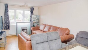 A living room with a brown leather sofa, a grey armchair, and a small table. There is a window with curtains, a fireplace, and a bookshelf.